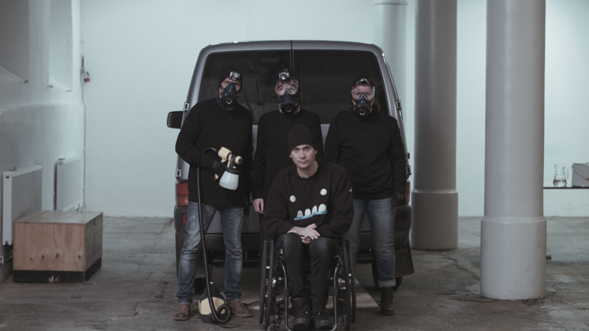 En skåpbil bakifrån, utanför står fyra svartklädda personer med gasmask och sprayfärg, en i rullstol