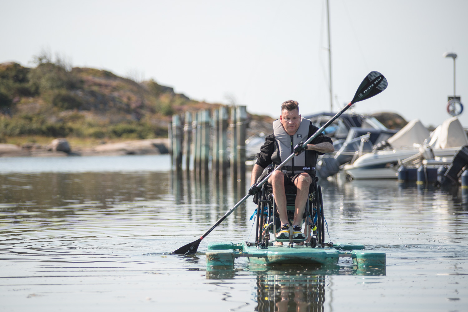 Person i rullstol på anpassad SUP, paddlar i småbåtshamn.