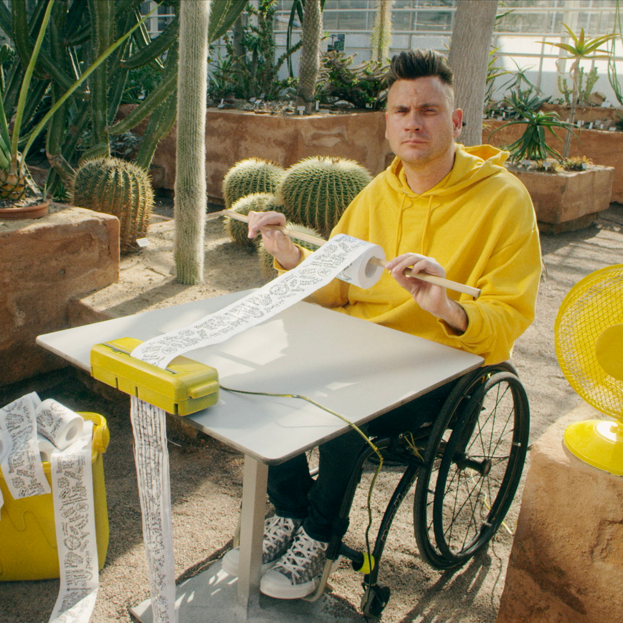 Man i rullstol, gul tröja, gul fläkt, gul dokumentförstörare, strimlar toalettpapper i ökenlik miljö med kaktusar.