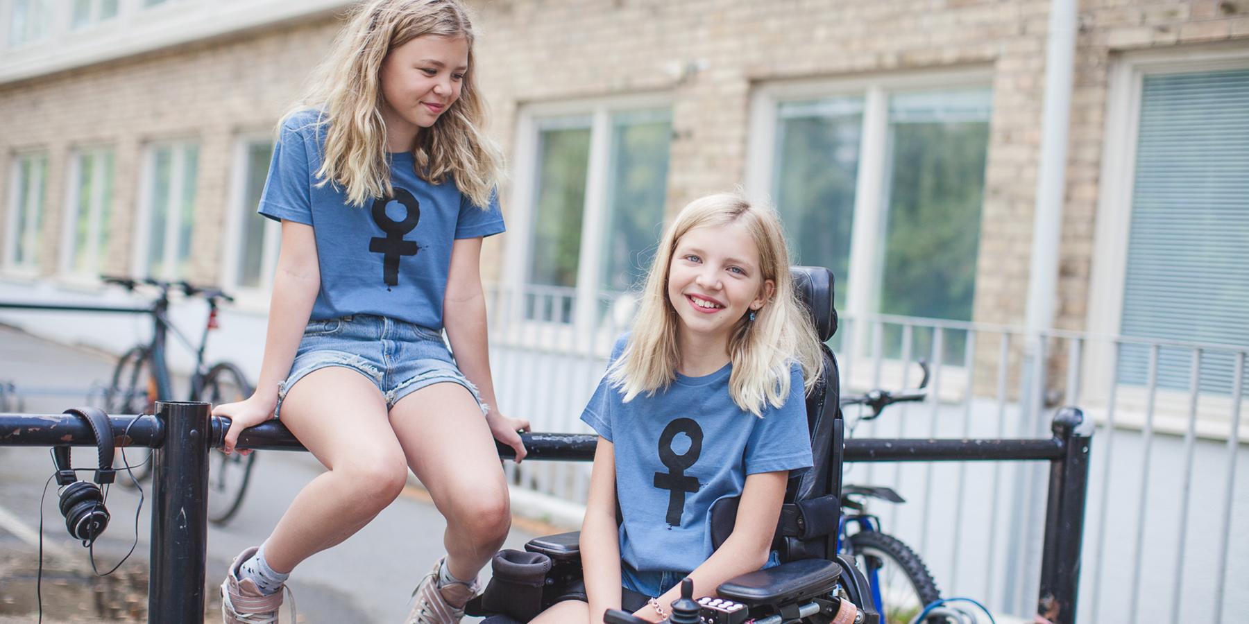 Två tjejer i t-shirts med kvinnosymboler på. Den ena tjejen sitter i elrullstol, den andra sitter på ett staket.