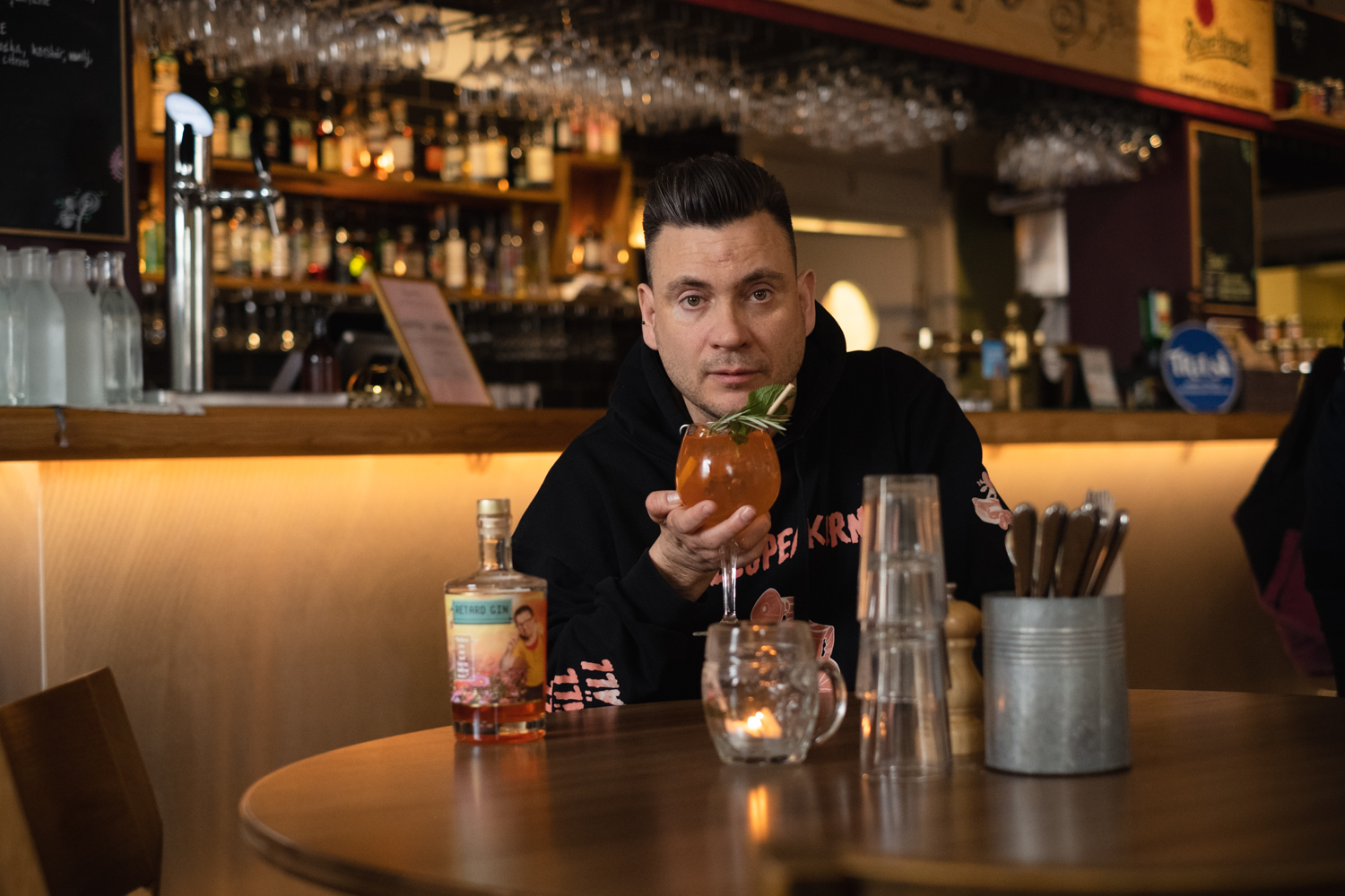 Anders Westgerd på en krog med en drink i handen och en flaska Retard Gin på bordet jämte sig.