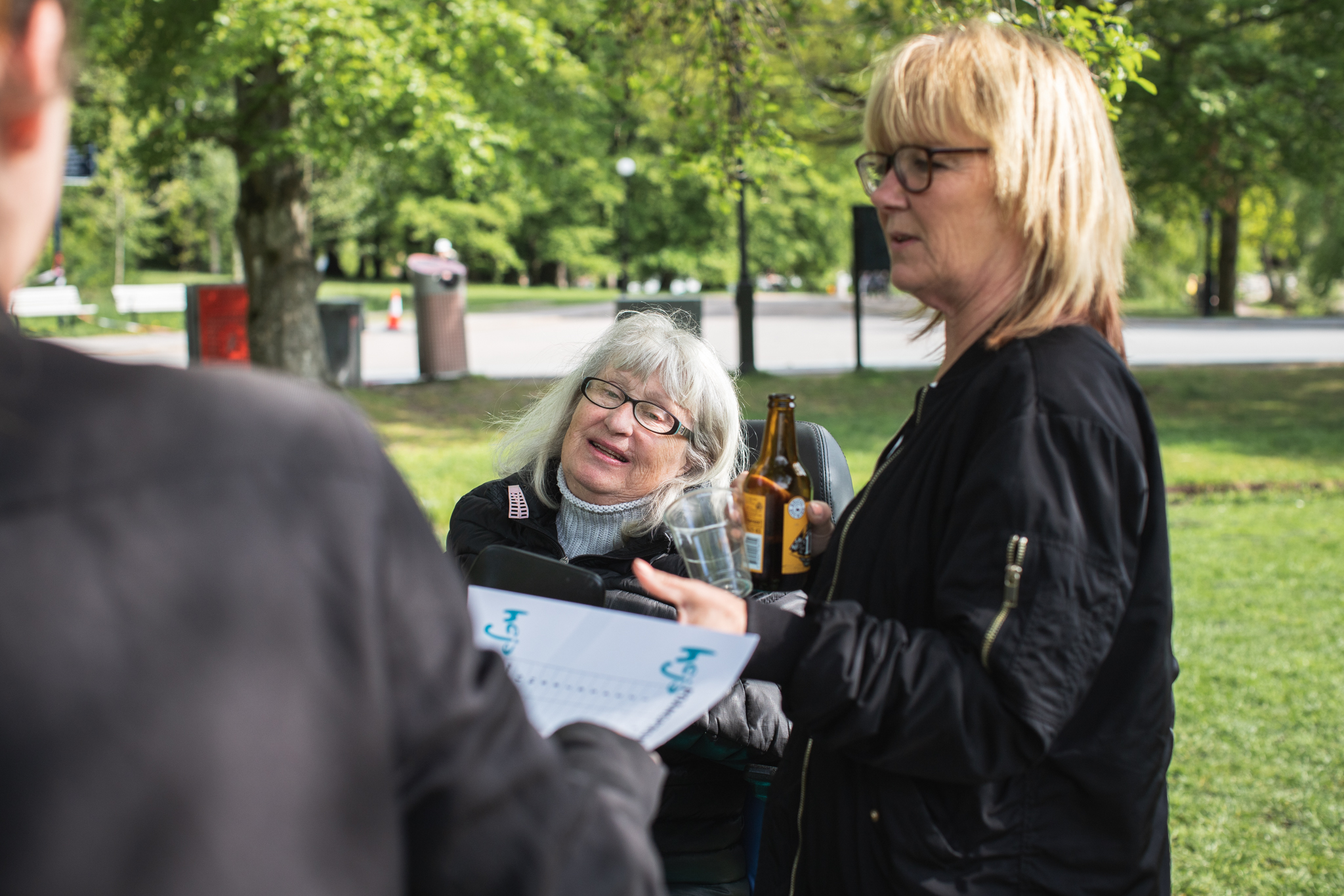 Kvinna i rullstol pratar med person som delar ut papper, en personlig assistent tar emot papperet, parkmiljö.