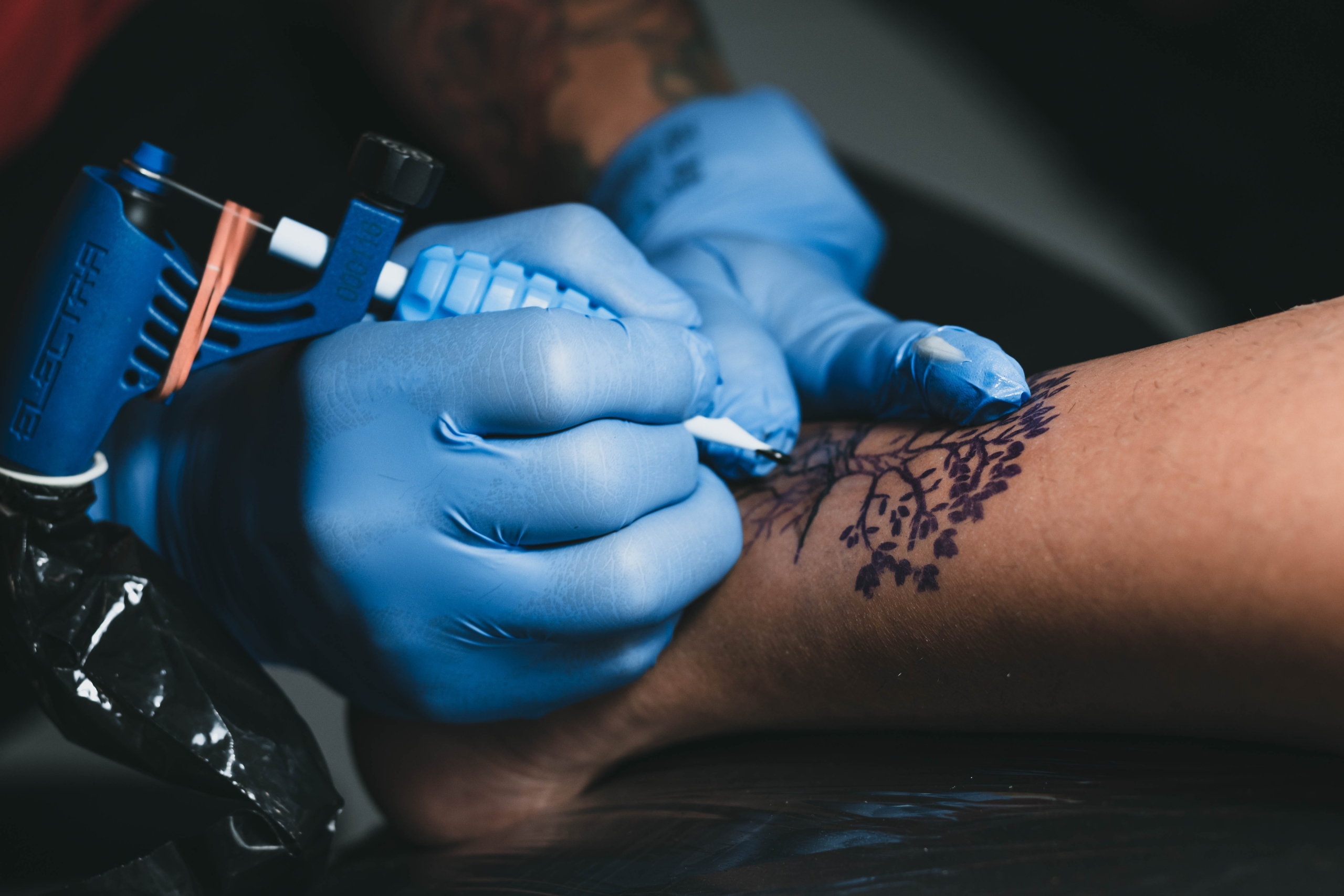 Bild på händer med blåa handskar och tatueringsmaskin, tatuerar ett träd
