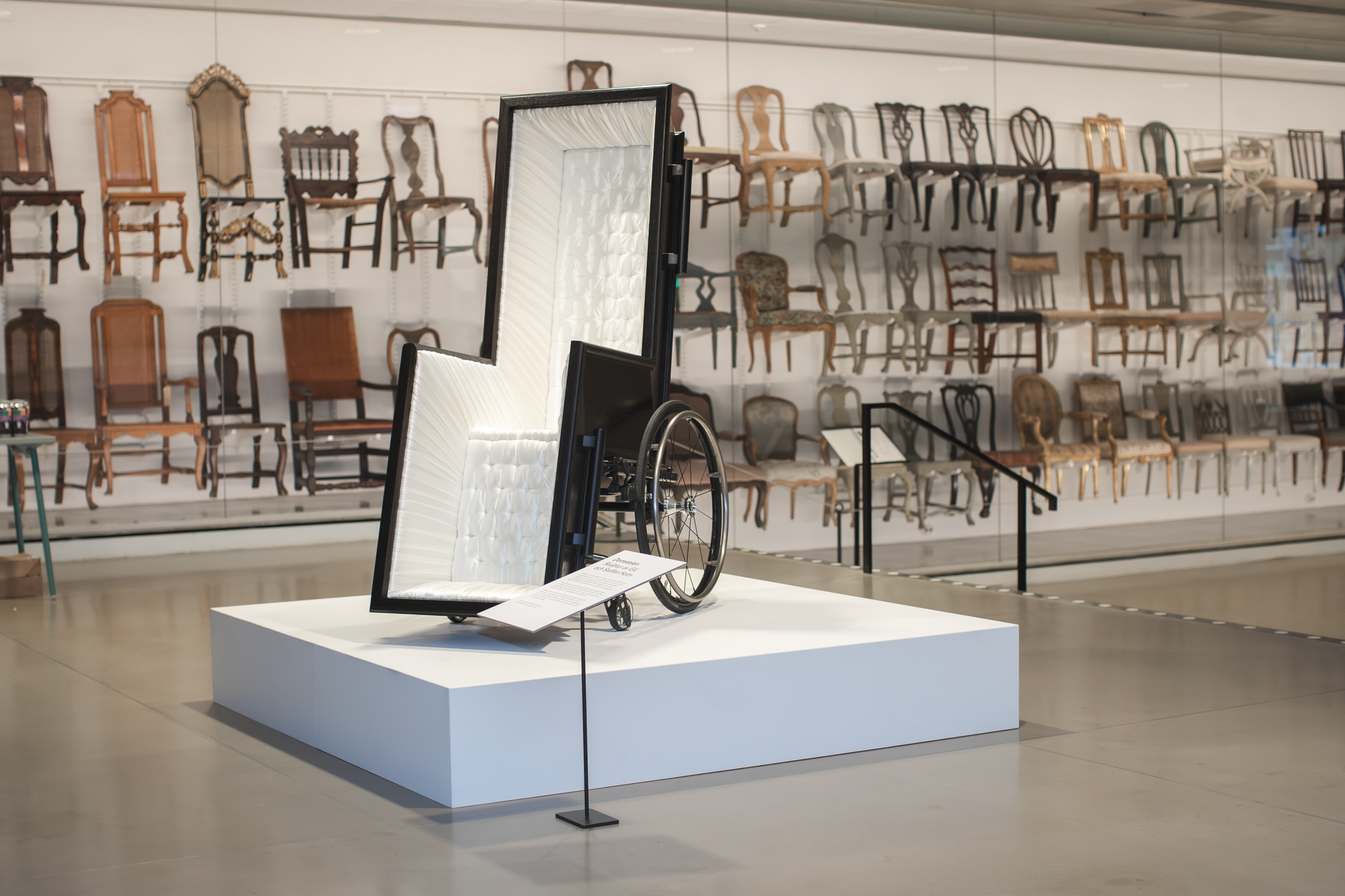 En likkista och en rullstol i ett, utställd på museum, en utställning med vanliga stolar syns i bakgrunden.