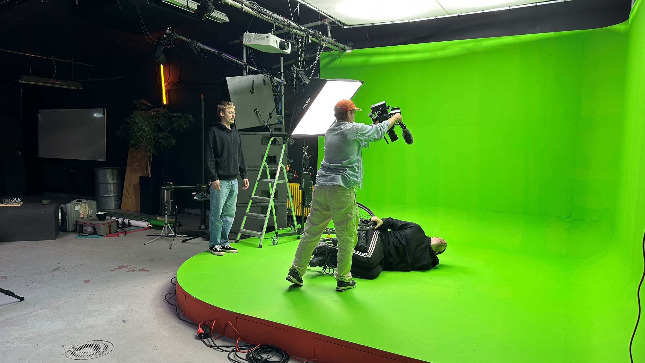 Behind the scenes-bild från filminspelning. Greenscreen, videografer och kameror, en person ligger på golvet i rullstol.
