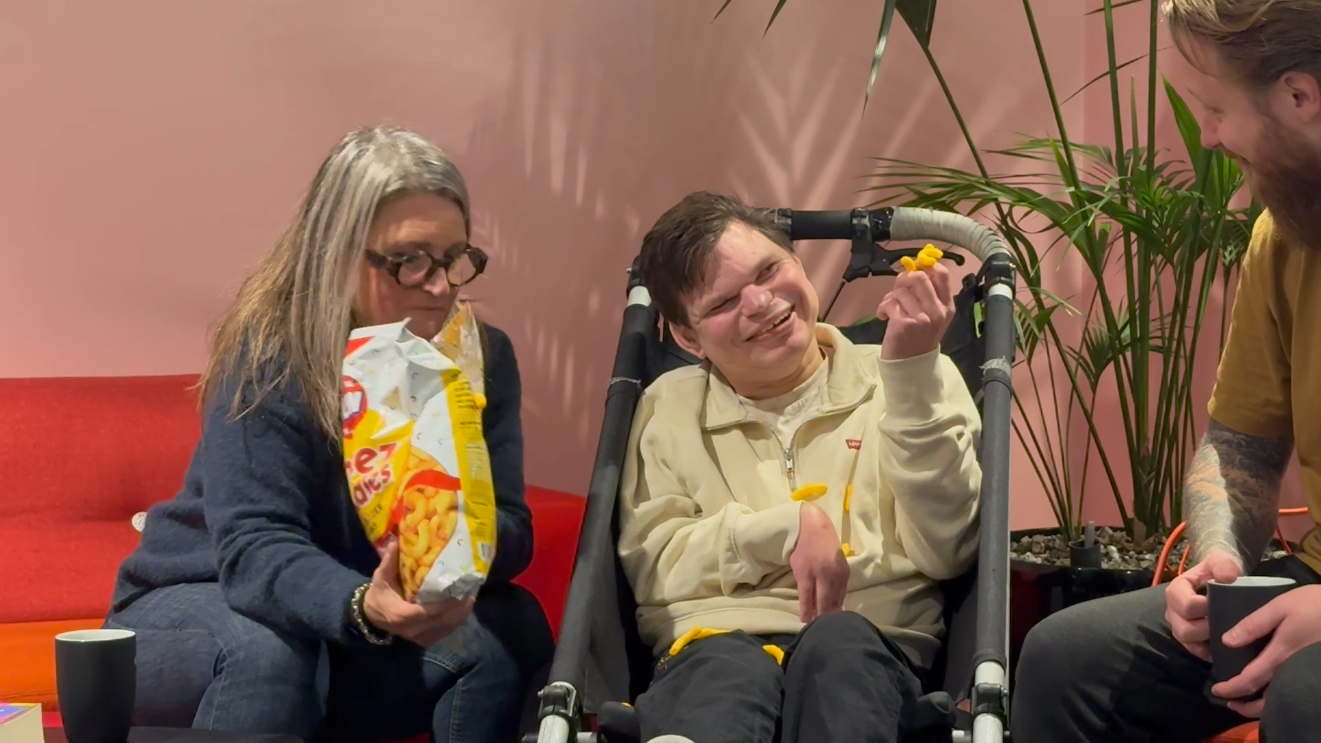 Kille i sulky äter ostbågar, hans mamma sitter jämte med påsen, killen ser glad ut.