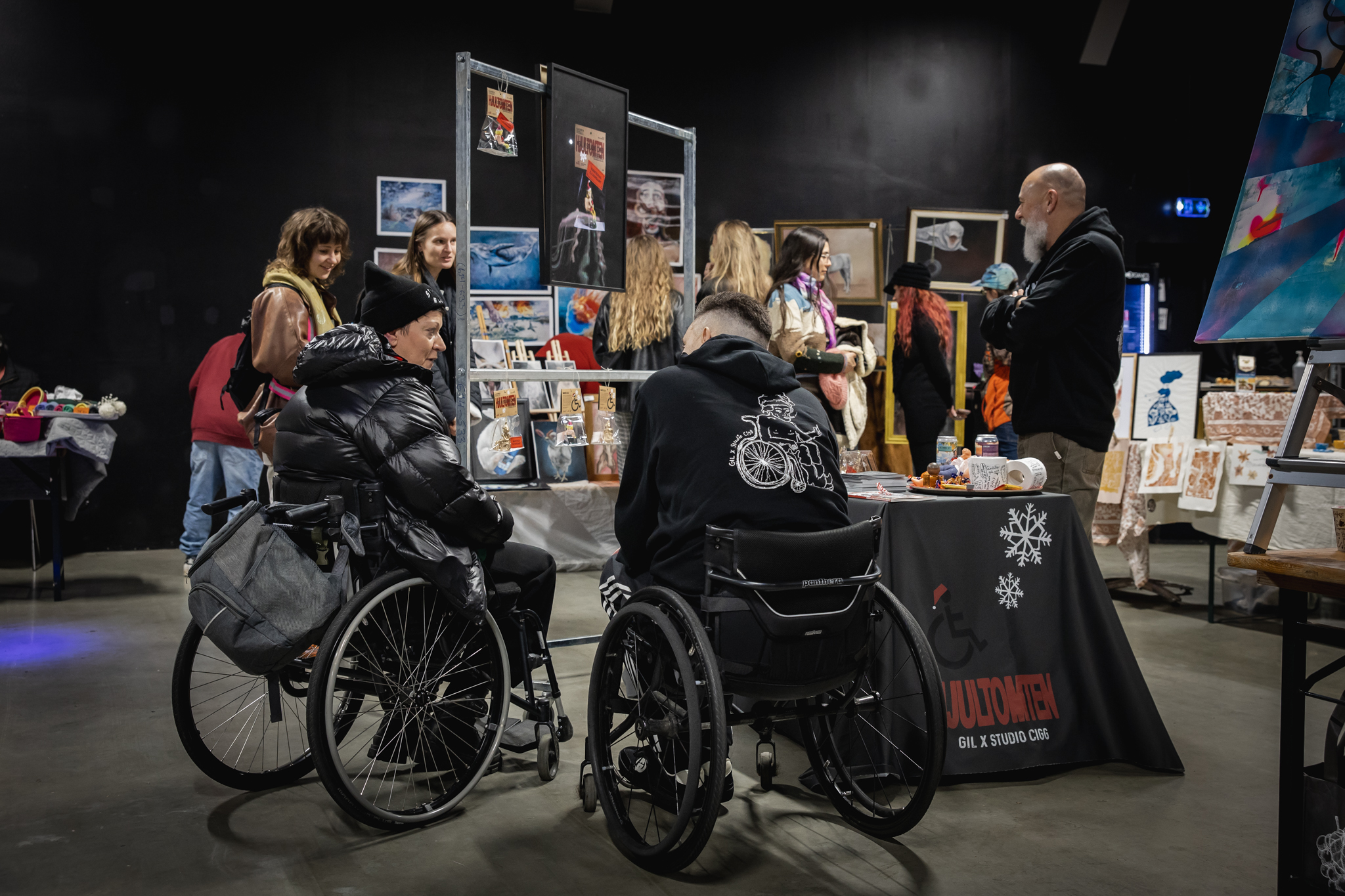 Från marknad, Ett antal personer samlas runt Hjultomten-montern, två sitter i rullstol. Svart a väggar, betonggolv, konst i bakgrunden.