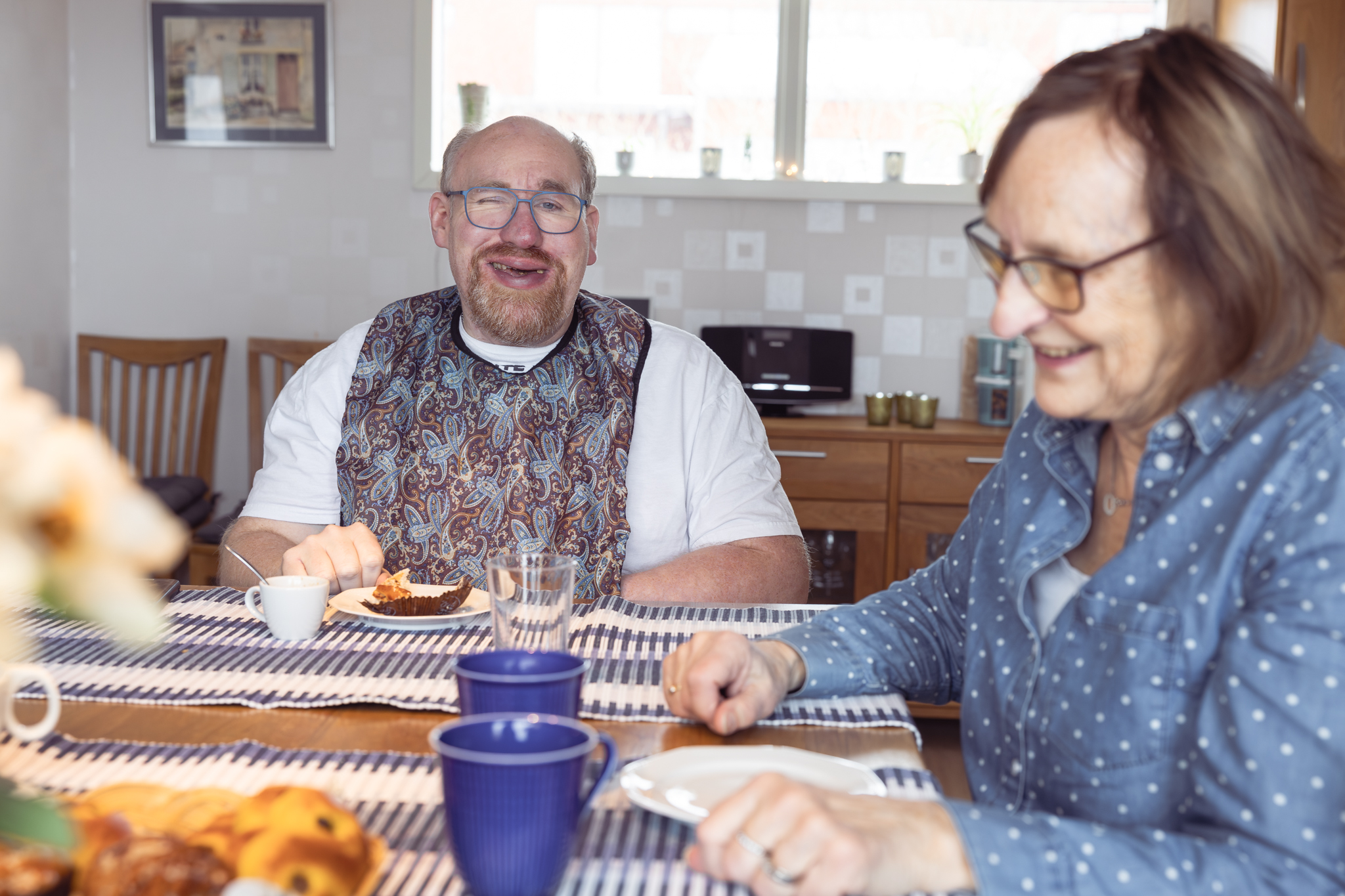 Matplatsmiljö, En man med hakklapp ler mot kameran, han har fika på ett fat framför sig, en  kvinna som kan vara hans mor i förgrnden, också hon är glad.