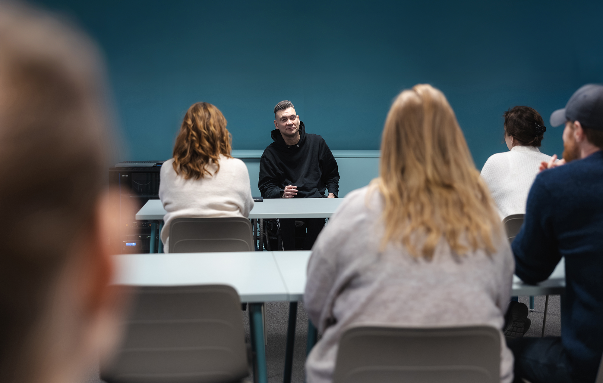 En föreläsning, man ser publiken bakifrån, deras hår och ryggar, längst bort i bild sitter föreläsaren, han sitter i rullstol.