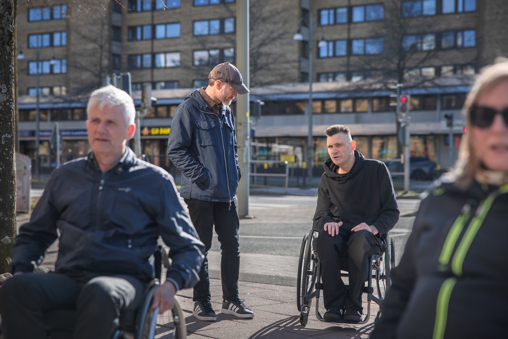 En man står och pratar med en annan man i rullstol. Två personer i rullstol i förgrunden. Stadsmiljö.