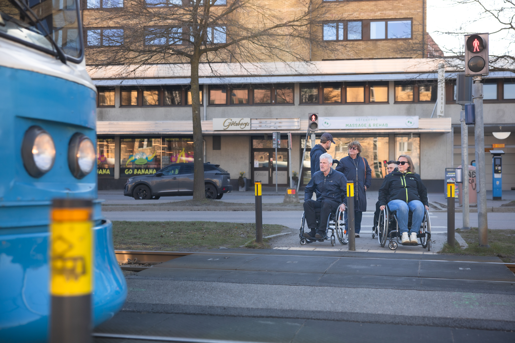 Ett sällskap med flera rullstolsburna och några gångare väntar vid rödljus, en spårvagn är på väg ut ur bild, stadsmiljö.