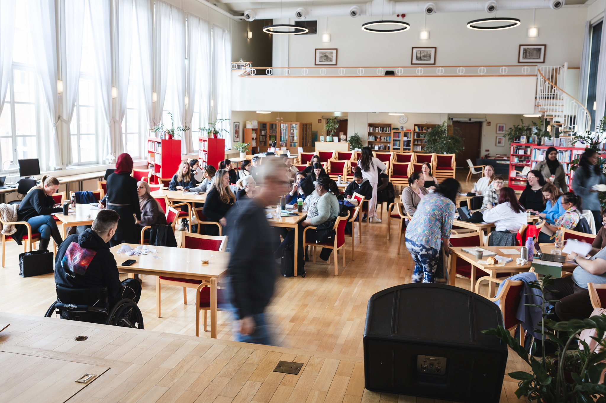 En föreläsningssal, bilden är tagen från scen. Närmst scenen sitter en man i rullstol, Det ser ut att vara paus, folk rör sig i lokalen som är ljus med parkettgolv och vita skira gardiner.