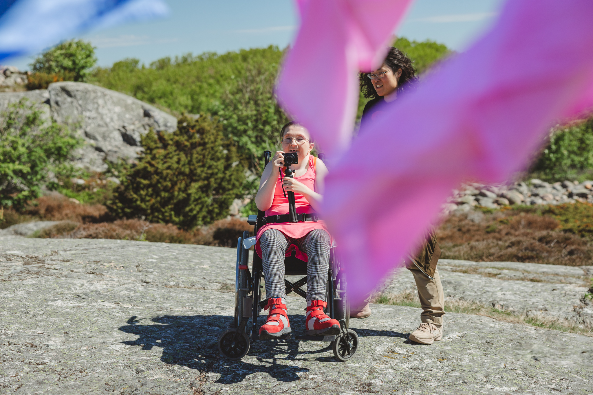 Rosa och blått tyg fladdrar framför kameran, mellan tygerna skymtas Anna i rullstol, som tar foton av tygerna.