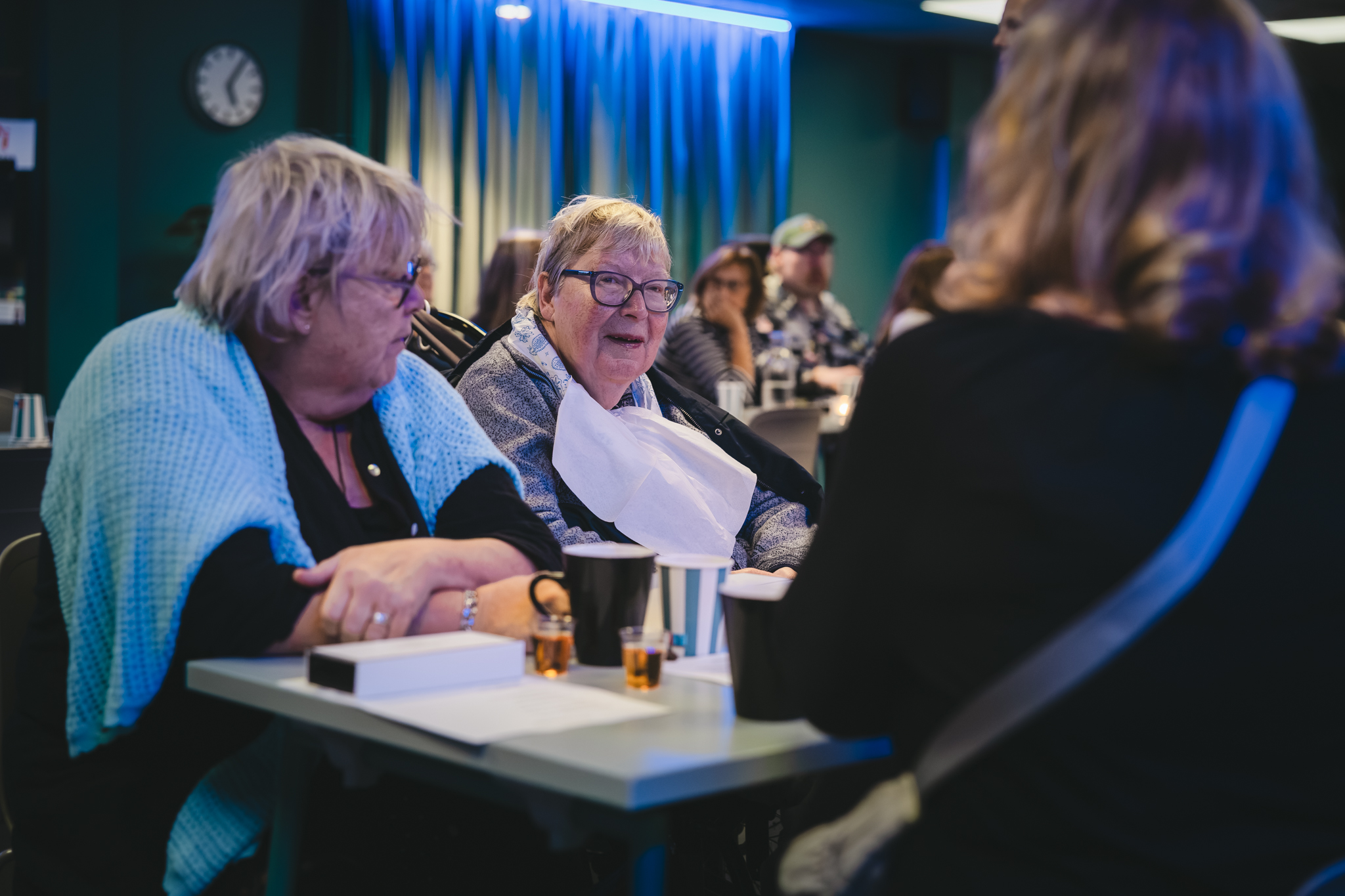 Tre kvinnor runt ett bord, de ser ut att prata med varandra. De sitter i ett rum med andra, väggarna är turkos och belysningen är neonblå.