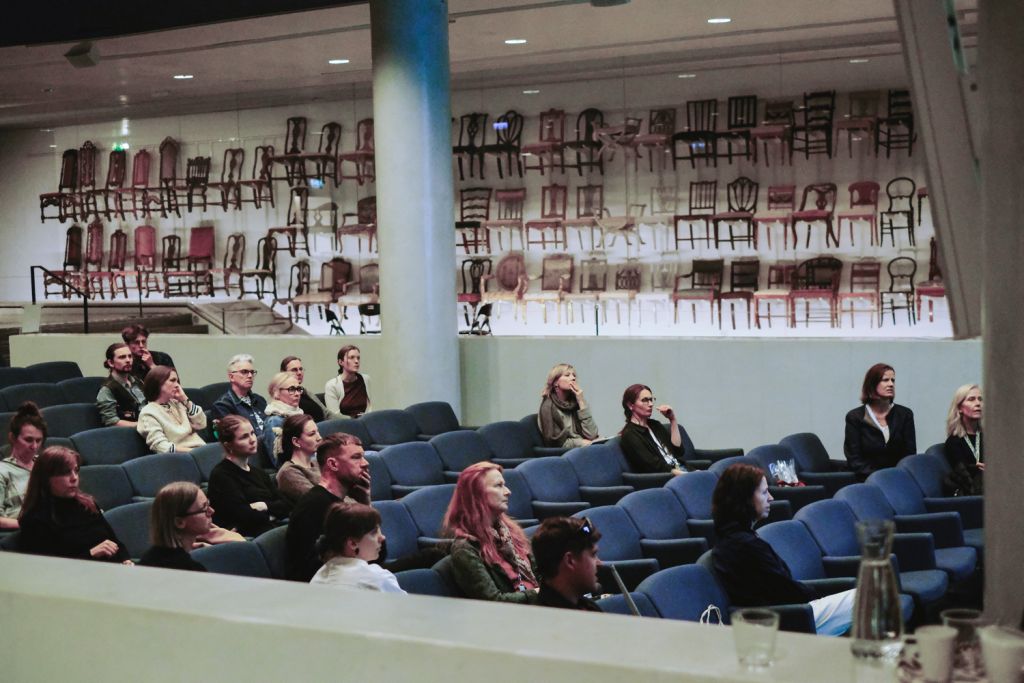 Publik som lyssnar til lnågon som man inte ser i bild, flera rader med blåa stolar. I bakgrunden en utställning med stolar längs med hela väggen.