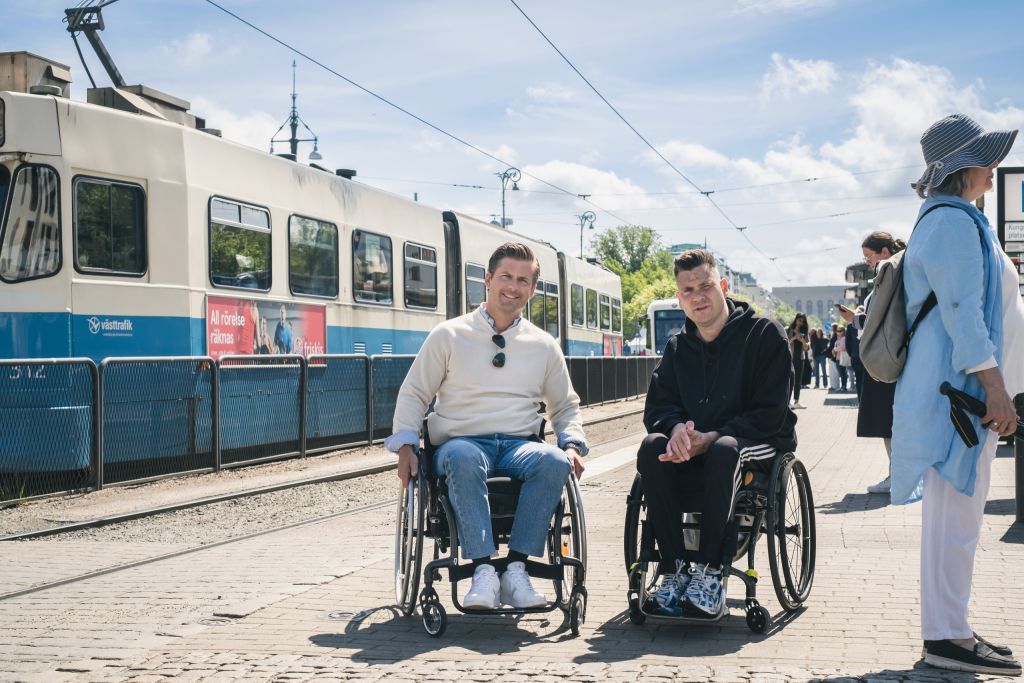 Hampus Magnuson och Anders Westgerd på spårvagnshållplats. Båda sitter i rullstol.